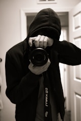 A person dressed in a dark hoodie holds a camera up to their face, capturing an image in a dimly lit room. The focus is on the camera lens, while the person's face is obscured by the hood and the camera. The environment seems to be indoors with visible doors in the background.