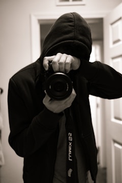 A person dressed in a dark hoodie holds a camera up to their face, capturing an image in a dimly lit room. The focus is on the camera lens, while the person's face is obscured by the hood and the camera. The environment seems to be indoors with visible doors in the background.