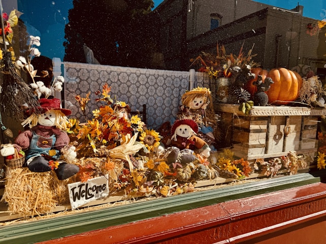 A window display featuring several scarecrow figures surrounded by autumn decorations including sunflowers, pumpkins, and leaves. A 'Welcome' sign is placed on a bale of hay, enhancing the festive fall theme. The setting has a warm, rustic feel with wooden elements and rich seasonal colors.