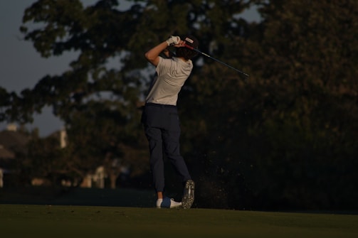 A golfer is captured mid-swing on a golf course, with their back to the camera. The player wears a white shirt and dark pants, holding a golf club in the air. The setting appears to be a lush, tree-lined area during a late afternoon or early evening, casting long shadows.