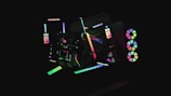A dark background highlights various abstract geometric shapes illuminated by neon colors. The shapes include circles, rectangles, and lines that create a sense of movement and modernity. Some elements resemble computer hardware with RGB lights, giving a futuristic aesthetic.