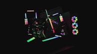 A dark background highlights various abstract geometric shapes illuminated by neon colors. The shapes include circles, rectangles, and lines that create a sense of movement and modernity. Some elements resemble computer hardware with RGB lights, giving a futuristic aesthetic.