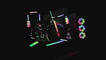 A dark background highlights various abstract geometric shapes illuminated by neon colors. The shapes include circles, rectangles, and lines that create a sense of movement and modernity. Some elements resemble computer hardware with RGB lights, giving a futuristic aesthetic.