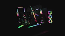 A dark background highlights various abstract geometric shapes illuminated by neon colors. The shapes include circles, rectangles, and lines that create a sense of movement and modernity. Some elements resemble computer hardware with RGB lights, giving a futuristic aesthetic.