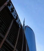 Tall skyscrapers with modern architectural design are seen against a clear blue sky, featuring glass facades. Prominent logos of large corporations appear on the buildings.
