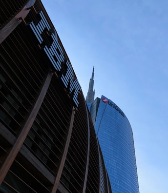 Tall skyscrapers with modern architectural design are seen against a clear blue sky, featuring glass facades. Prominent logos of large corporations appear on the buildings.