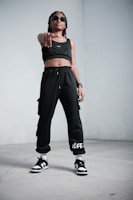 A person stands confidently indoors, wearing black athletic clothing including a crop top and sweatpants. They are also wearing sunglasses, sneakers, and have a watch on their wrist. The stance is assertive, with one hand pointing forward.