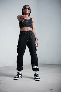 A person stands confidently indoors, wearing black athletic clothing including a crop top and sweatpants. They are also wearing sunglasses, sneakers, and have a watch on their wrist. The stance is assertive, with one hand pointing forward.