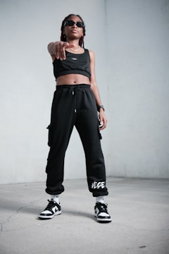 A person stands confidently indoors, wearing black athletic clothing including a crop top and sweatpants. They are also wearing sunglasses, sneakers, and have a watch on their wrist. The stance is assertive, with one hand pointing forward.