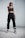 A person stands confidently indoors, wearing black athletic clothing including a crop top and sweatpants. They are also wearing sunglasses, sneakers, and have a watch on their wrist. The stance is assertive, with one hand pointing forward.