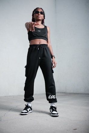 A person stands confidently indoors, wearing black athletic clothing including a crop top and sweatpants. They are also wearing sunglasses, sneakers, and have a watch on their wrist. The stance is assertive, with one hand pointing forward.