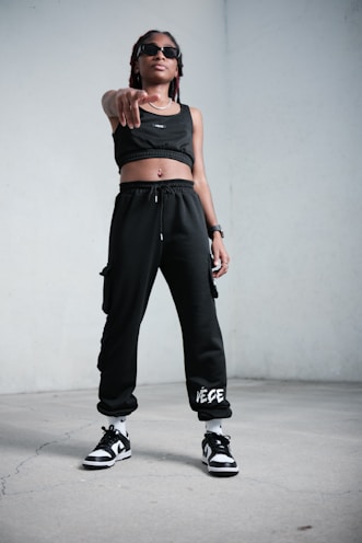 A person stands confidently indoors, wearing black athletic clothing including a crop top and sweatpants. They are also wearing sunglasses, sneakers, and have a watch on their wrist. The stance is assertive, with one hand pointing forward.