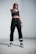 A person stands confidently indoors, wearing black athletic clothing including a crop top and sweatpants. They are also wearing sunglasses, sneakers, and have a watch on their wrist. The stance is assertive, with one hand pointing forward.