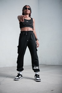 A person stands confidently indoors, wearing black athletic clothing including a crop top and sweatpants. They are also wearing sunglasses, sneakers, and have a watch on their wrist. The stance is assertive, with one hand pointing forward.