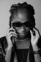A person wearing sunglasses holds the frames with both hands. The individual has braided hair and is wearing a necklace with star-shaped pendants. The attire includes a leather jacket.