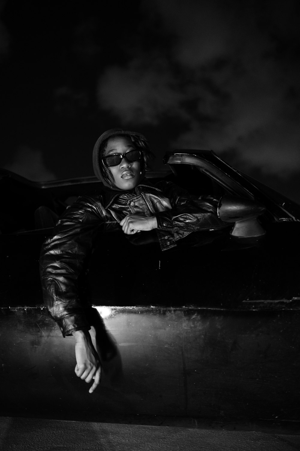 a black and white photo of a man leaning against a car