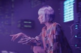 A person with short, silver hair is depicted in a futuristic setting with dim purple lighting. They are wearing a brightly colored, patterned outfit with elongated sleeves. The background features various electronic or digital components, as well as bright, illuminated screens with unreadable text or symbols.
