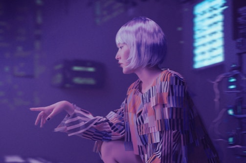 A person with short, silver hair is depicted in a futuristic setting with dim purple lighting. They are wearing a brightly colored, patterned outfit with elongated sleeves. The background features various electronic or digital components, as well as bright, illuminated screens with unreadable text or symbols.