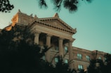 An elegant university building featuring classical architectural elements such as ornate columns and a detailed pediment. The building is partially obscured by trees in the foreground, and the sky is a clear blue, casting a warm glow over the structure.