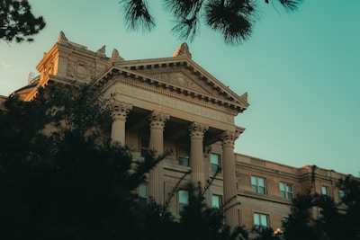 An elegant university building featuring classical architectural elements such as ornate columns and a detailed pediment. The building is partially obscured by trees in the foreground, and the sky is a clear blue, casting a warm glow over the structure.
