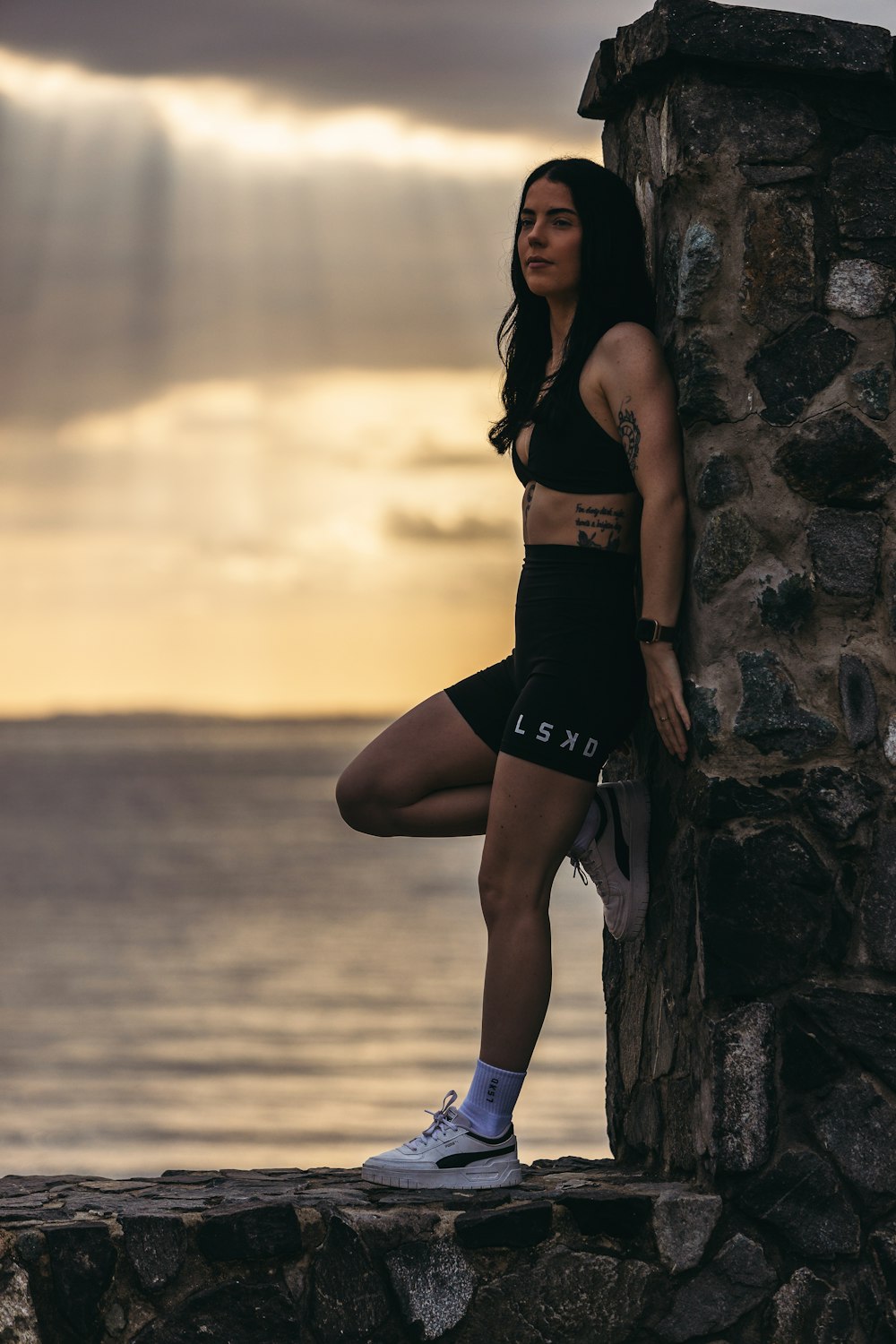 a woman leaning against a stone wall near the ocean