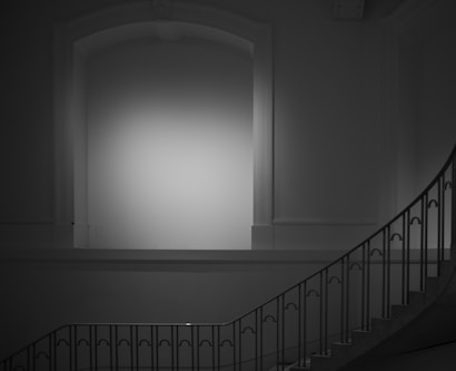 A monochromatic image featuring a minimalist interior space with a large, empty wall in the background. The room includes an elegant, curved staircase with a simple metal railing on the side. The lighting casts a soft glow on the wall, highlighting its architectural details.