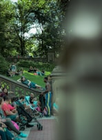 A lively outdoor scene in a park featuring several people sitting and interacting on stone steps and benches. A man in athletic wear stands by a group, engaging in conversation. The lush greenery of trees provides a natural canopy in the background.