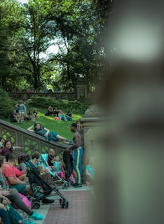 A lively outdoor scene in a park featuring several people sitting and interacting on stone steps and benches. A man in athletic wear stands by a group, engaging in conversation. The lush greenery of trees provides a natural canopy in the background.