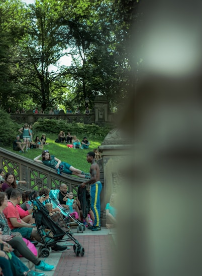 A lively outdoor scene in a park featuring several people sitting and interacting on stone steps and benches. A man in athletic wear stands by a group, engaging in conversation. The lush greenery of trees provides a natural canopy in the background.