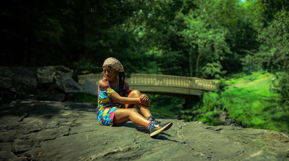 a young girl sitting on a rock with a bridge in the background