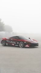 A sleek red sports car with black and white detailing is parked on a foggy road. The car is adorned with various logos and branding including 'NXT LEVEL AUTO PROTECTION'. There are blurry trees in the background, contributing to a serene and mystical atmosphere.