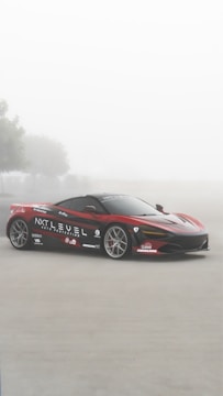 A sleek red sports car with black and white detailing is parked on a foggy road. The car is adorned with various logos and branding including 'NXT LEVEL AUTO PROTECTION'. There are blurry trees in the background, contributing to a serene and mystical atmosphere.