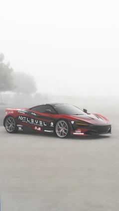 A sleek red sports car with black and white detailing is parked on a foggy road. The car is adorned with various logos and branding including 'NXT LEVEL AUTO PROTECTION'. There are blurry trees in the background, contributing to a serene and mystical atmosphere.