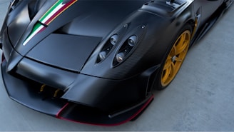 A sleek sports car features a carbon fiber body with Italian flag stripes on the hood. The headlight design is prominent, and the wheels are a striking gold color, set against a gray pavement background.