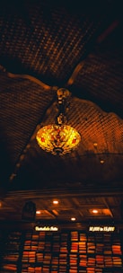 A decorative ceiling light with a stained glass design hangs in a dimly lit room. The ceiling is crafted with dark, intricately curved wooden beams. Below, shelves are stacked with neatly arranged vibrant fabrics illuminated by warm, soft lights, with signs indicating price ranges.