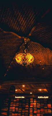 A decorative ceiling light with a stained glass design hangs in a dimly lit room. The ceiling is crafted with dark, intricately curved wooden beams. Below, shelves are stacked with neatly arranged vibrant fabrics illuminated by warm, soft lights, with signs indicating price ranges.