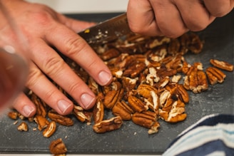 A person is using a knife to chop pecans on a cutting board. There are many nuts scattered across the board, indicating active food preparation. One hand is holding the knife while the other is steadying the nuts.