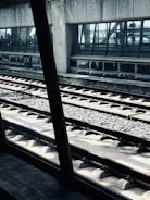 A railway platform with tracks running parallel in the foreground. A large glass window reveals a waiting area inside a building, where silhouettes of people can be seen. The concrete walls and the metal structures offer an industrial and modern architectural style. Sunlight casts shadows and highlights parts of the tracks, giving a dynamic visual effect.