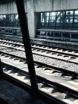A railway platform with tracks running parallel in the foreground. A large glass window reveals a waiting area inside a building, where silhouettes of people can be seen. The concrete walls and the metal structures offer an industrial and modern architectural style. Sunlight casts shadows and highlights parts of the tracks, giving a dynamic visual effect.