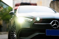 A sleek, modern car with a prominent front grille featuring a silver logo. Bright headlights are illuminated, and the car's design exudes luxury and sophistication. The background is blurred, highlighting the vehicle's details.