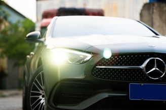 A sleek, modern car with a prominent front grille featuring a silver logo. Bright headlights are illuminated, and the car's design exudes luxury and sophistication. The background is blurred, highlighting the vehicle's details.