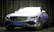 A luxury car with a sleek, modern design is parked indoors. The vehicle has a metallic body with a prominent grille and stylized headlights. The environment is dimly lit with textured dark curtains in the background, creating a sophisticated setting.