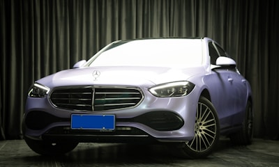 A luxury car with a sleek, modern design is parked indoors. The vehicle has a metallic body with a prominent grille and stylized headlights. The environment is dimly lit with textured dark curtains in the background, creating a sophisticated setting.
