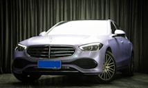 A luxury car with a sleek, modern design is parked indoors. The vehicle has a metallic body with a prominent grille and stylized headlights. The environment is dimly lit with textured dark curtains in the background, creating a sophisticated setting.