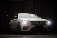 A luxurious white car is parked indoors against a dark curtain backdrop. The car's headlights are on, and a lens flare is visible above the vehicle. The car's sleek and modern design is highlighted, with emphasis on its distinctive front grille and emblem.