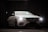 A luxurious white car is parked indoors against a dark curtain backdrop. The car's headlights are on, and a lens flare is visible above the vehicle. The car's sleek and modern design is highlighted, with emphasis on its distinctive front grille and emblem.