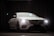 A luxurious white car is parked indoors against a dark curtain backdrop. The car's headlights are on, and a lens flare is visible above the vehicle. The car's sleek and modern design is highlighted, with emphasis on its distinctive front grille and emblem.