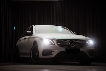 A luxurious white car is parked indoors against a dark curtain backdrop. The car's headlights are on, and a lens flare is visible above the vehicle. The car's sleek and modern design is highlighted, with emphasis on its distinctive front grille and emblem.
