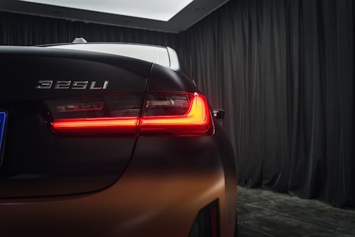 A rear view of a car with a model designation badge '325Li' displayed, showing a modern tail light design illuminated in vibrant red. The vehicle is positioned indoors with a backdrop of dark black curtains that create a sleek and elegant ambiance.