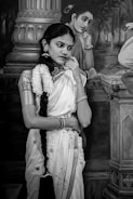A woman in traditional attire stands pensively in front of a painting. She is adorned with jewelry and a long braid decorated with flowers. The background features a classical-style painted image of another woman in a similar pose.